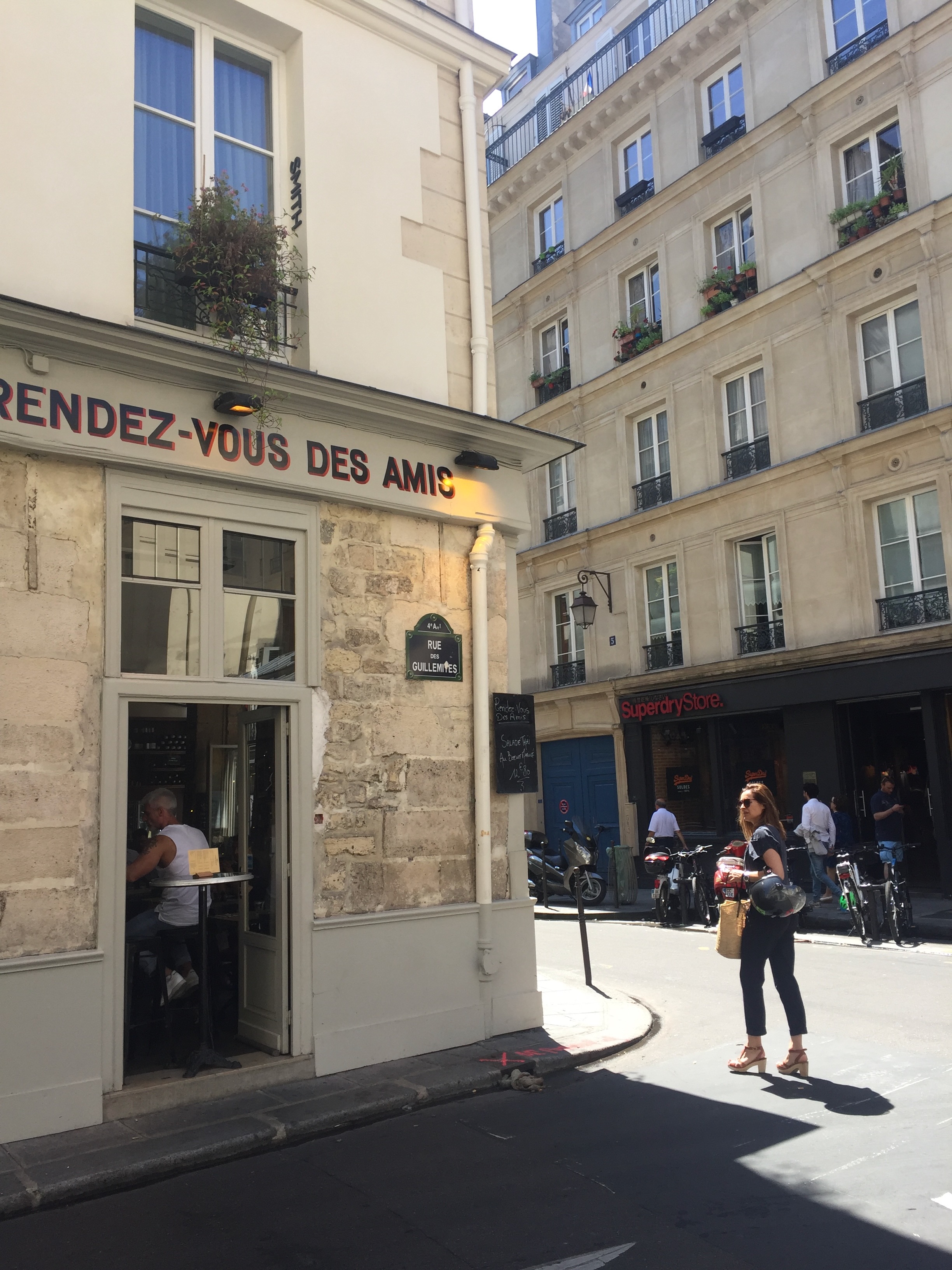 Street view in Paris with a café named "Rendez-vous des Amis" on a sunny day. A woman walks by carrying a helmet and a bag, with buildings in the background.