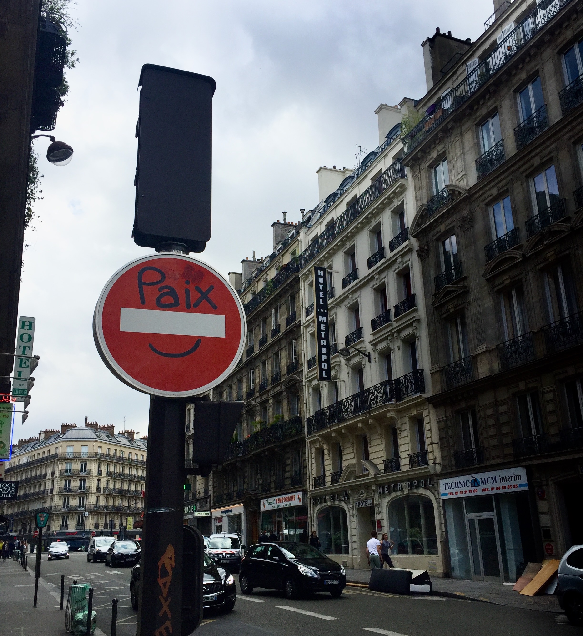 A creative "Do Not Enter" sign in Paris with "Paix" (peace) and a smiley face drawn on it. Overcast sky, classic Parisian buildings, street view.